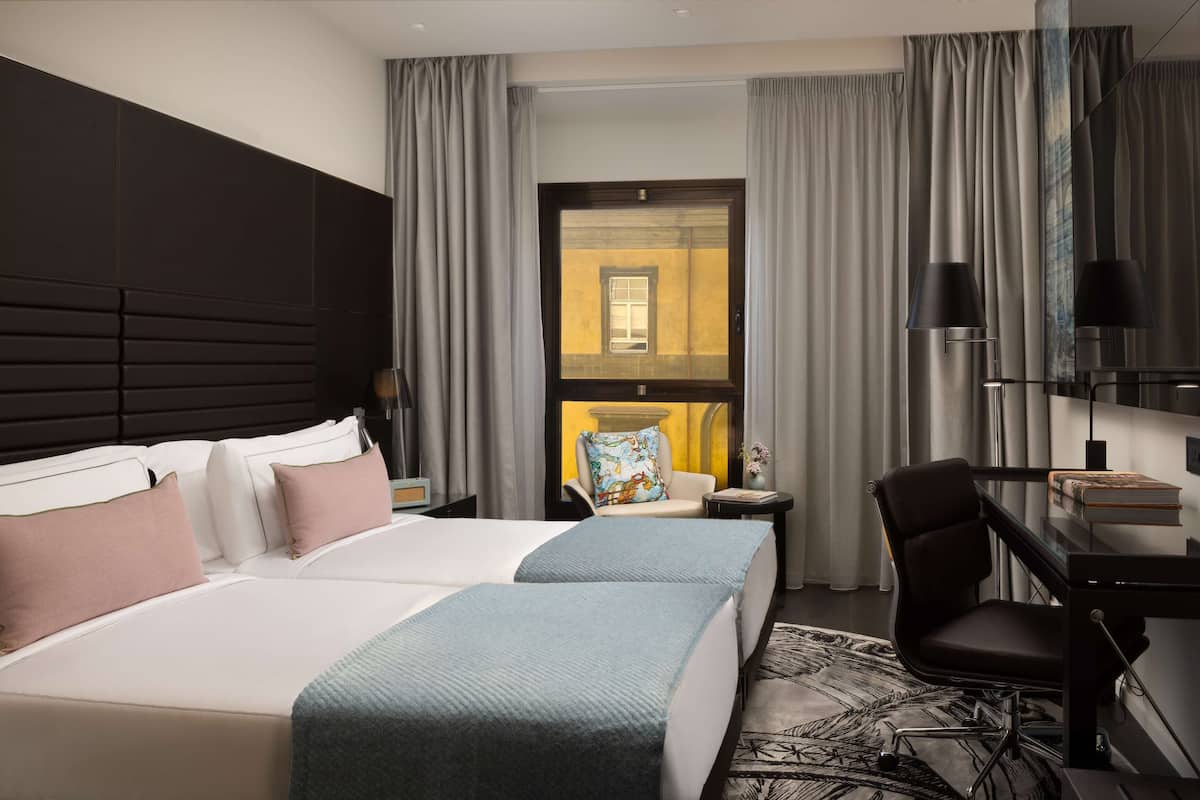 A hotel room with two beds, a desk, a chair, and a view of a building through a window.