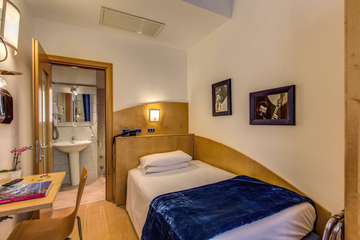 A hotel room with a bed, a desk with a chair, a sink, and a mirror.