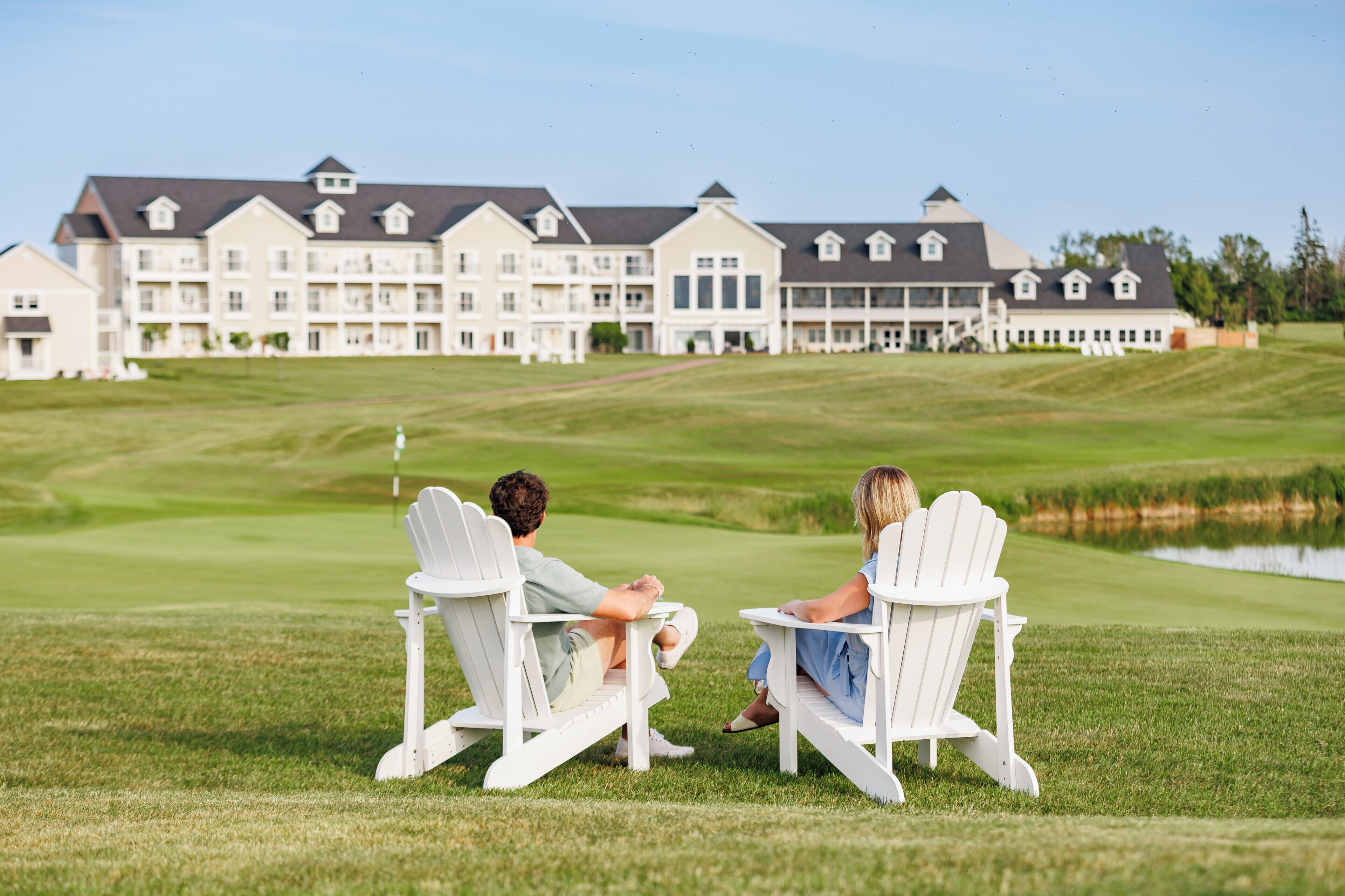 Two people seated on Adirondack chairs facing a golf course with a large building in the background.