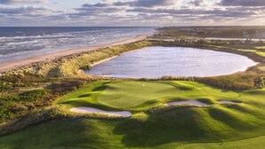 Un parcours de golf doté d’un grand obstacle d’eau, d’une plage de sable et offrant une vue sur l’océan.