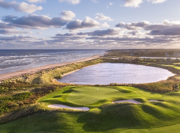 A golf course with a large water hazard, a sandy beach, and a view of the ocean.