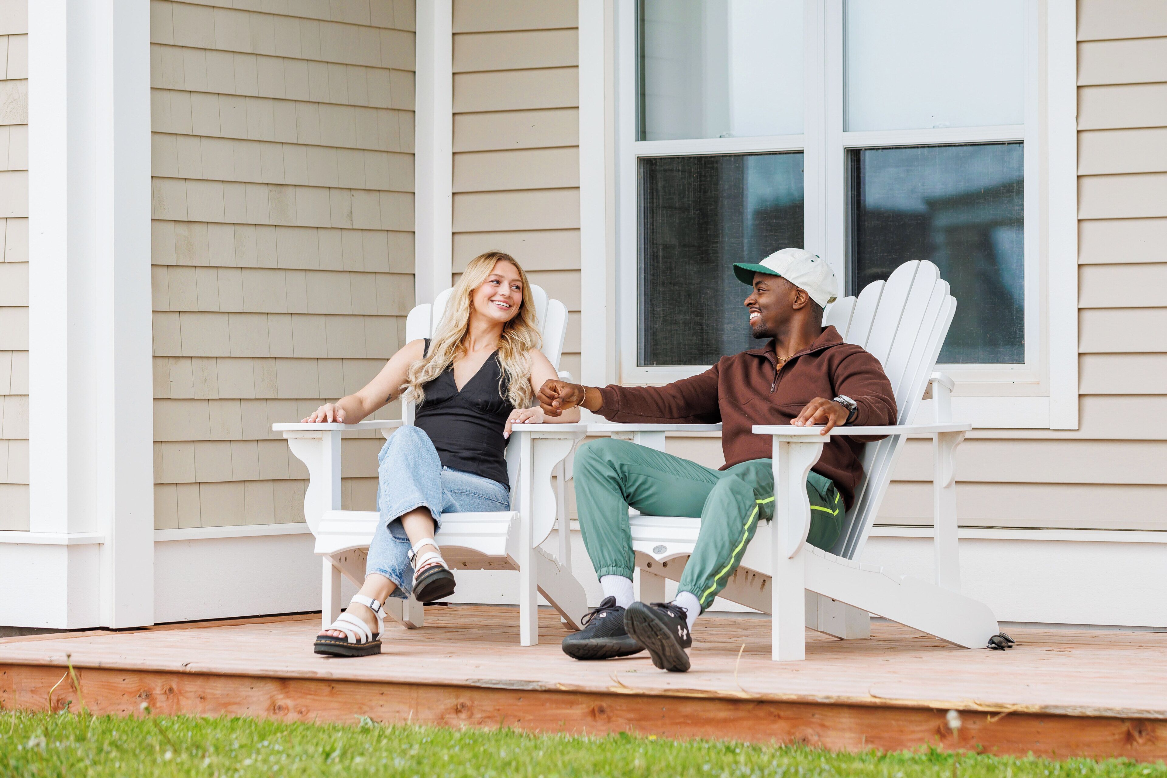 Dos personas sentadas en sillas Adirondack blancas sobre una terraza de madera frente a una casa con revestimiento color beige y un ventanal grande.