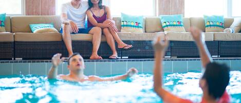 Zona de piscina con una pareja sentada en un sofá y gente nadando.