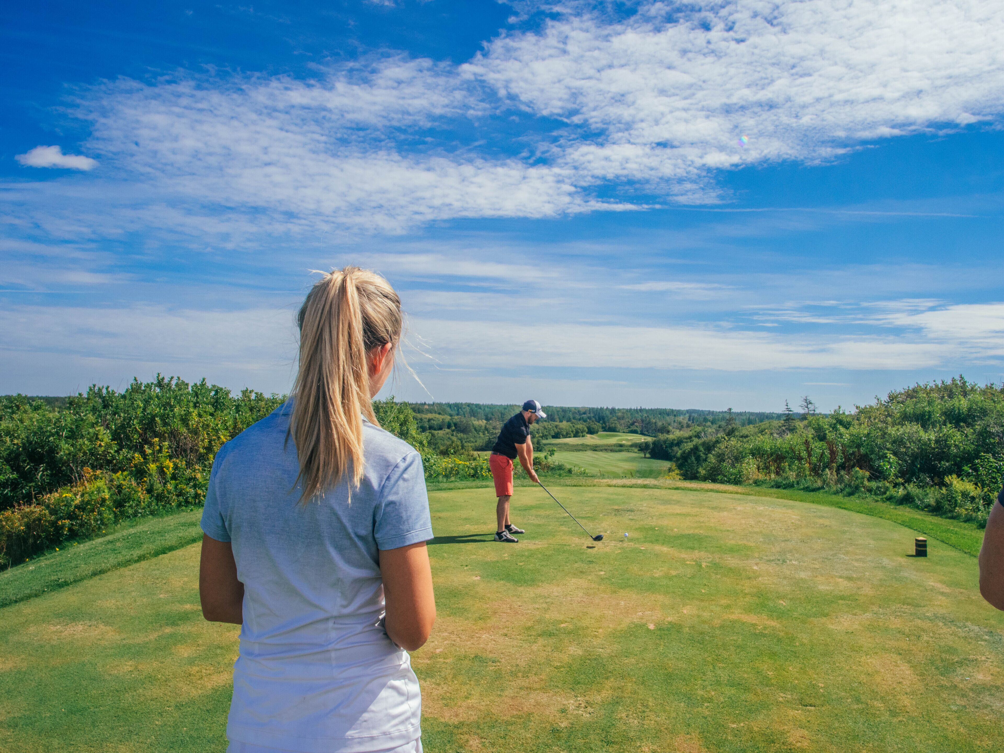 Un terrain de golf sous un ciel dégagé, un golfeur au loin et une personne debout sur l’herbe.
