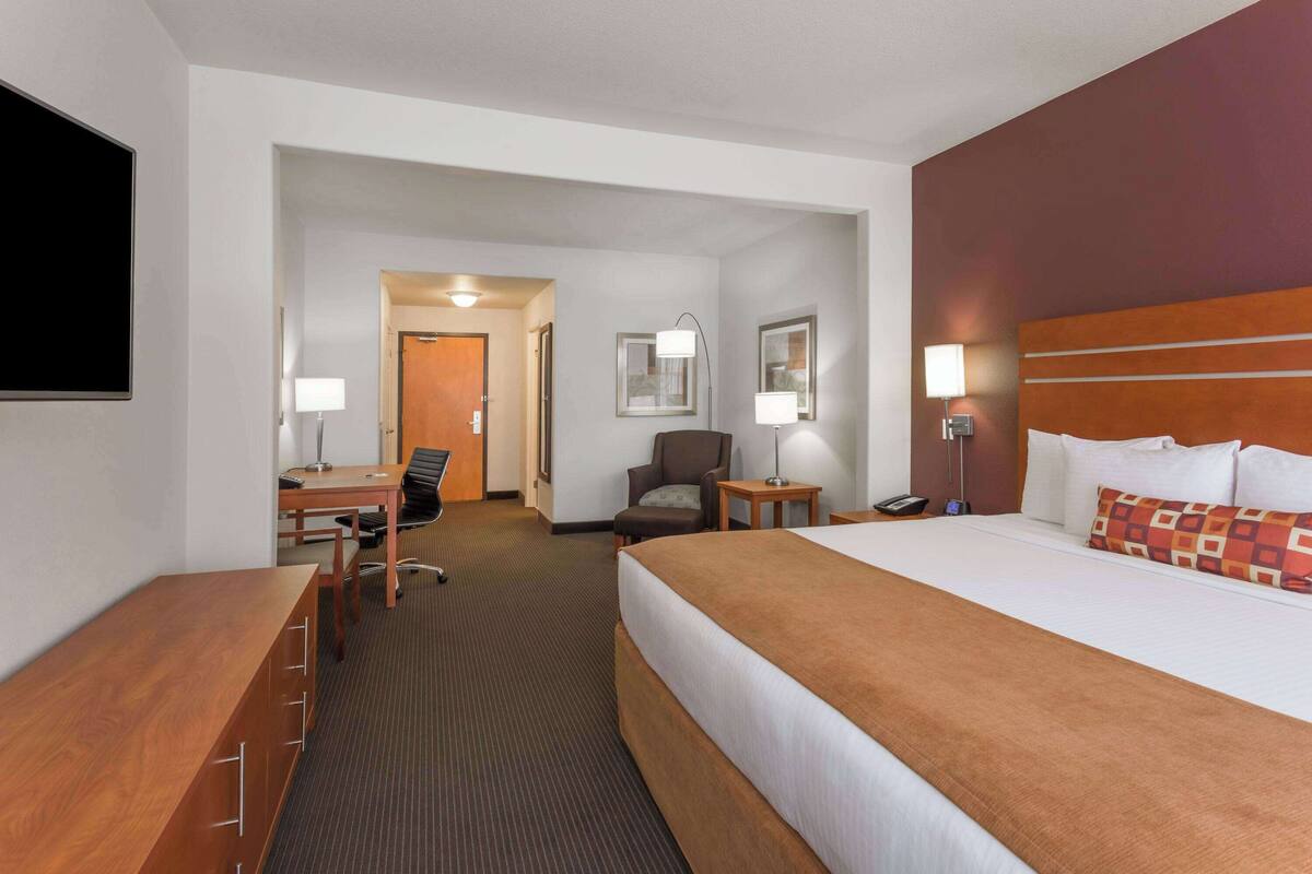 A hotel room with a large bed, a desk with a chair, a small table, and a lamp.