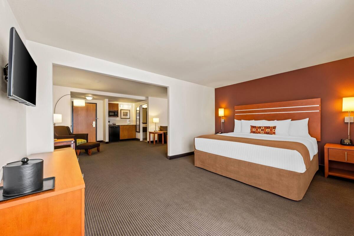 A hotel room with a large bed, a desk with a lamp, a television, and a view of the hallway and kitchen area.
