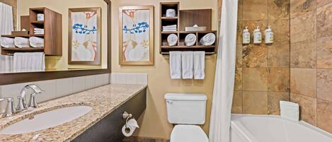 Un baño con lavamanos, inodoro y bañera. Hay toallas y artículos de aseo en estantes.