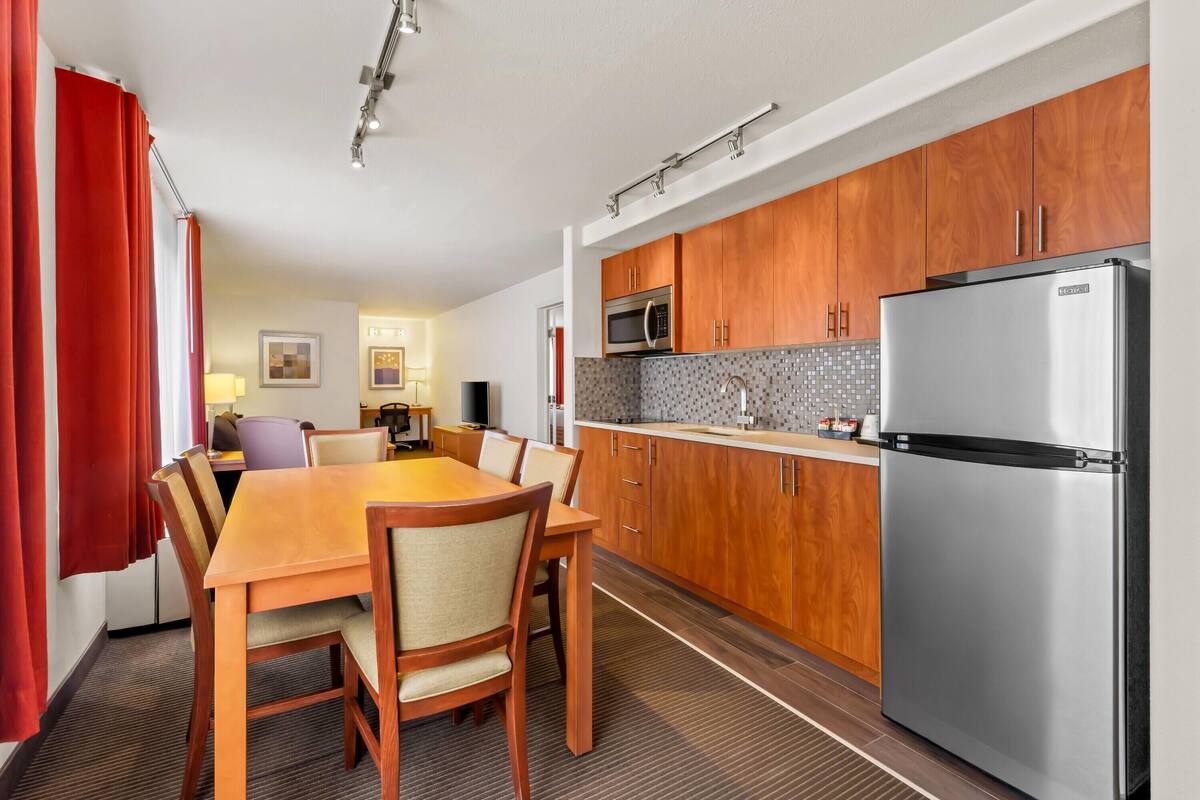 A modern kitchen with wooden cabinets, a stainless steel refrigerator, and a dining area with a wooden table and chairs.