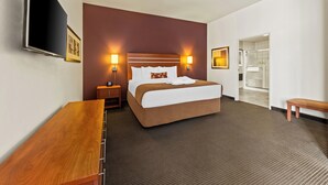 Habitación de hotel con una cama grande, lámparas de noche, un televisor de pantalla plana, un banco de madera y un baño visible a través de una puerta abierta.