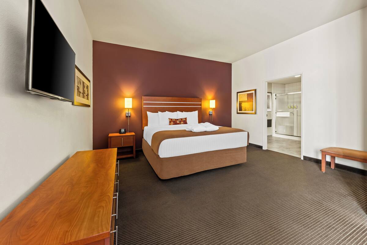 A hotel room with a large bed, bedside lamps, a flat-screen TV, a wooden bench, and a bathroom visible through an open door.