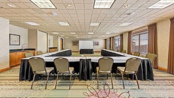 Un salón de conferencias con una larga mesa, sillas, una pantalla de proyector y un ventanal con persianas.