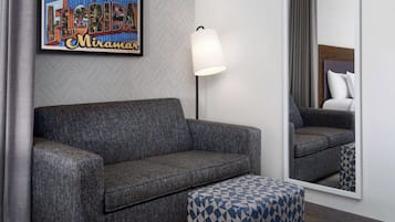 Une chambre d’hôtel avec un canapé gris, un pouf à motifs, un panneau encadré avec l’inscription « Salutations de Floride », un lampadaire et un grand miroir qui reflète la pièce.