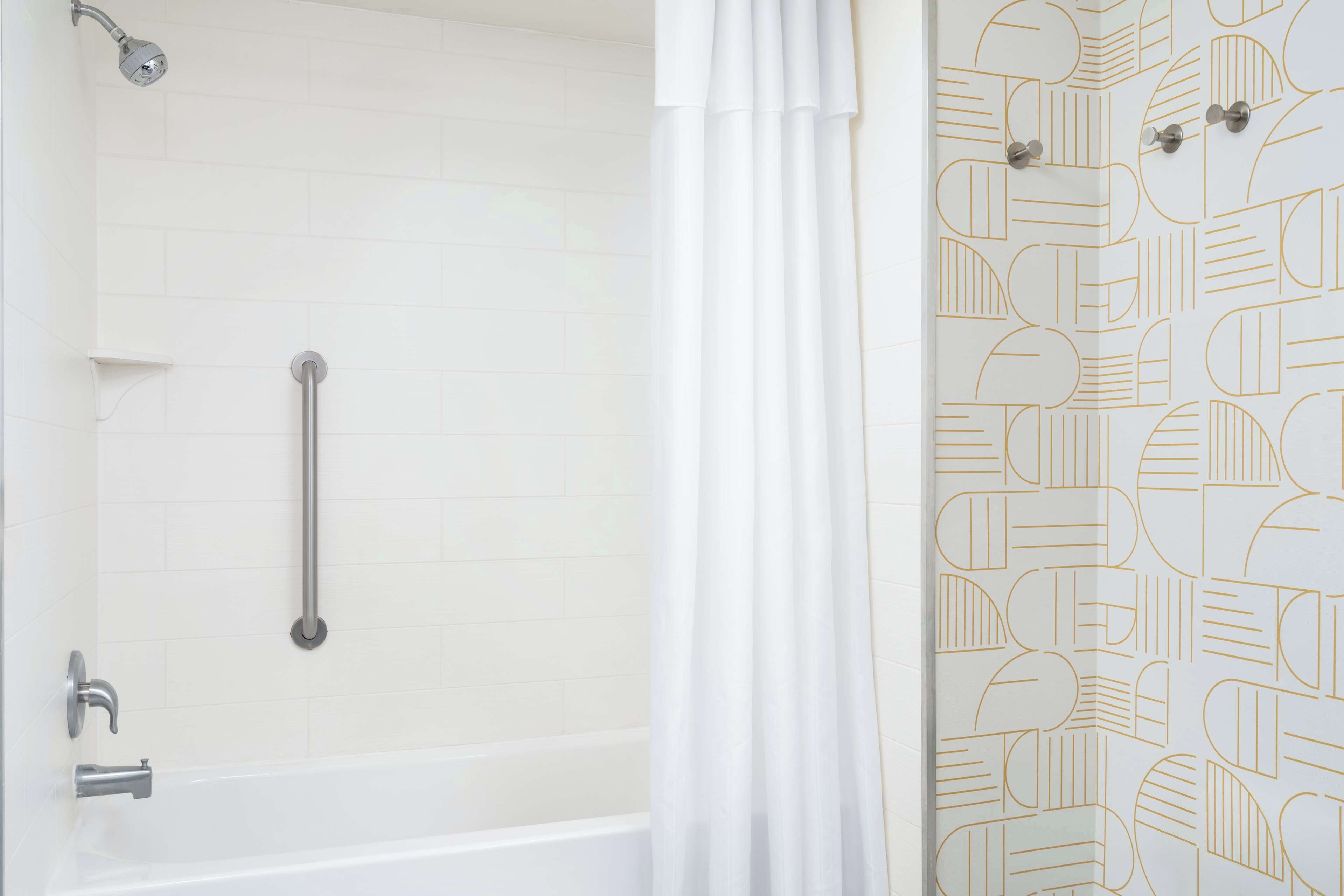 Un baño con pared de azulejos blancos, una bañera, una ducha con cabezal de ducha manual y una cortina blanca.