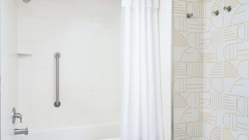 Un baño con pared de azulejos blancos, una bañera, una ducha con cabezal de ducha manual y una cortina blanca.