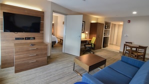 A modern hotel room with a flat-screen TV mounted on a wooden panel, a wooden dresser with drawers, a blue sofa, a small wooden table, and a kitchenette area in the background. - Holiday Inn Express & Suites Indianapolis Northwest by IHG (Indianapolis)