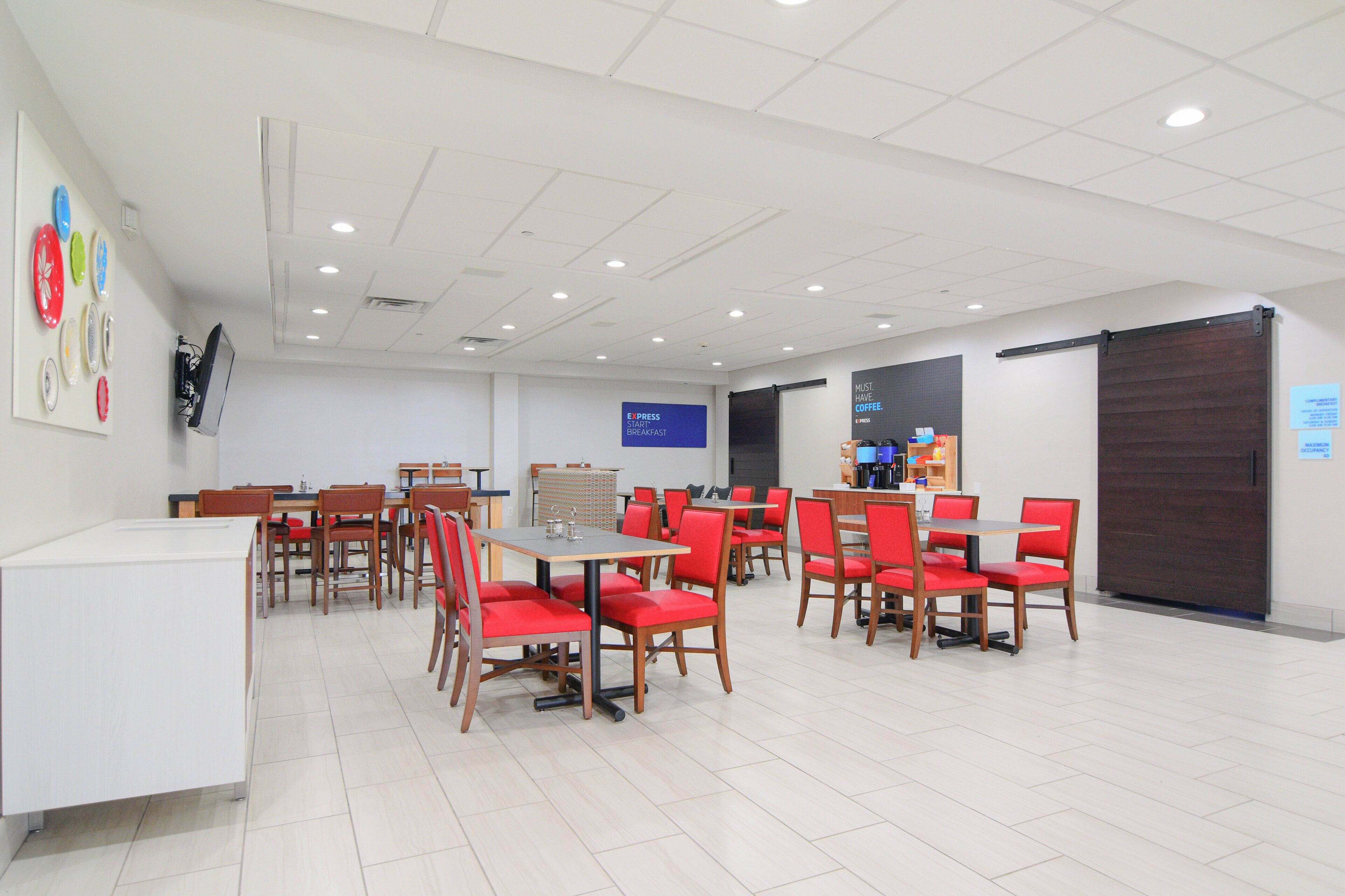 A spacious dining area with multiple tables and chairs, a coffee station, and a bulletin board with a sign.