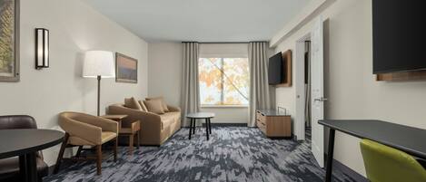 A hotel room with a sofa, chairs, a small table, a lamp, a TV, and a window with curtains.