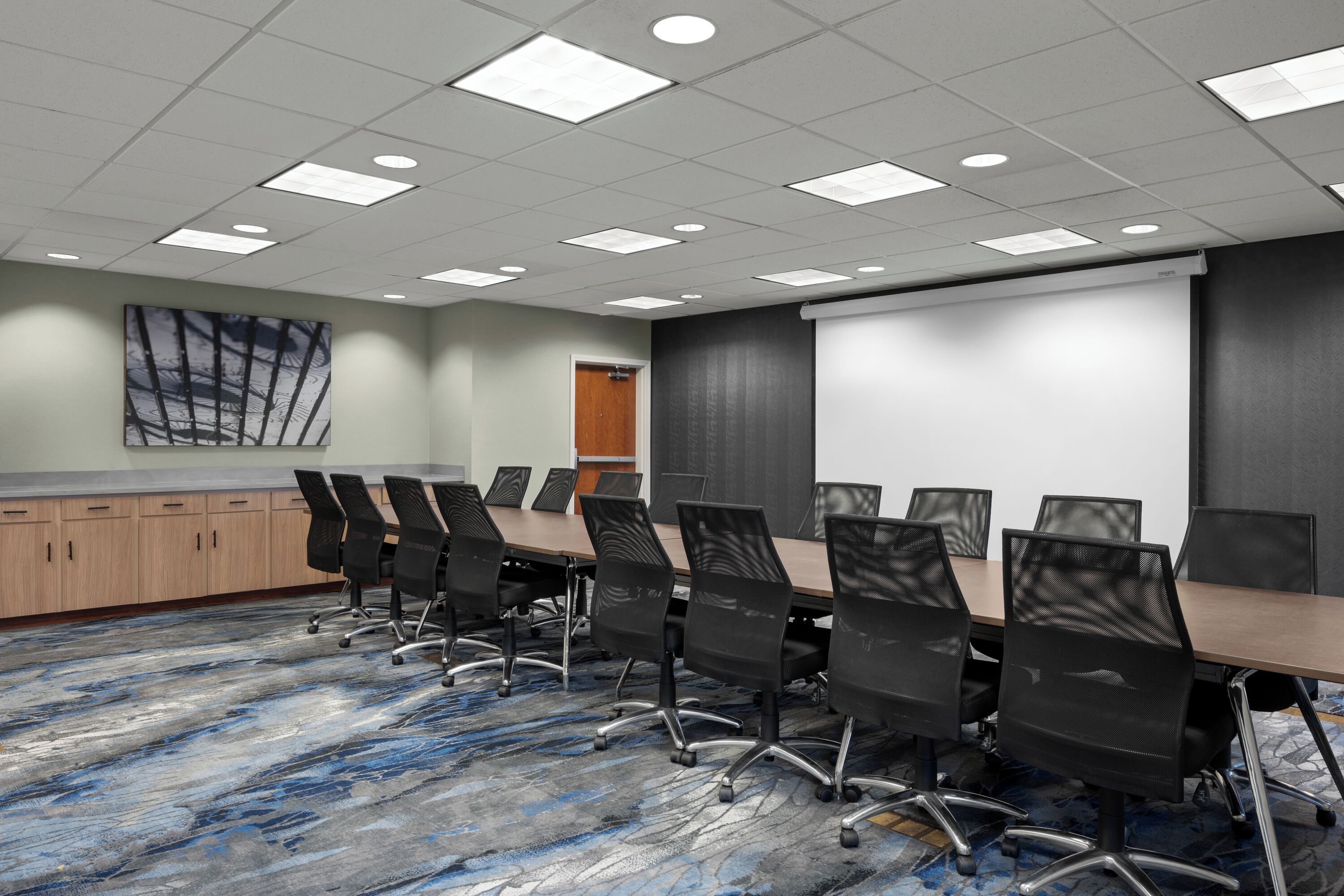 A conference room with a long table, black chairs, a projector screen, and a carpeted floor.