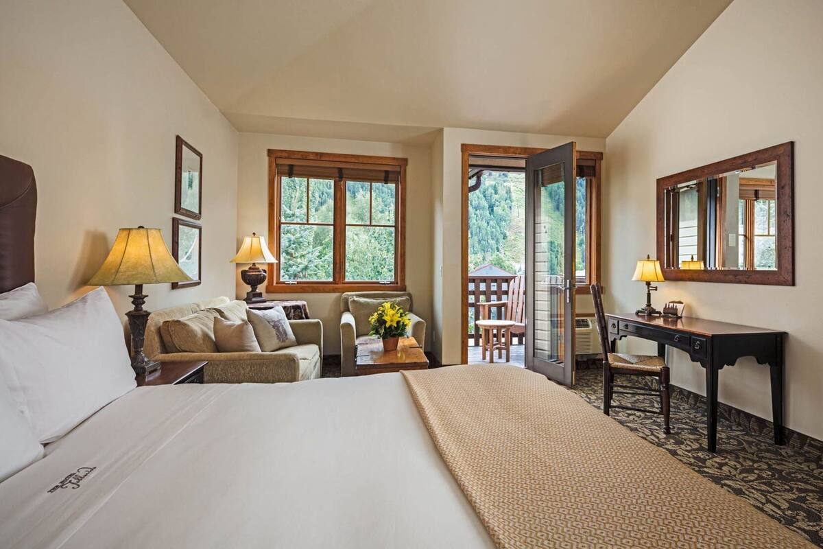 A hotel room with a bed, two chairs, a desk, a lamp, a window, and a balcony.