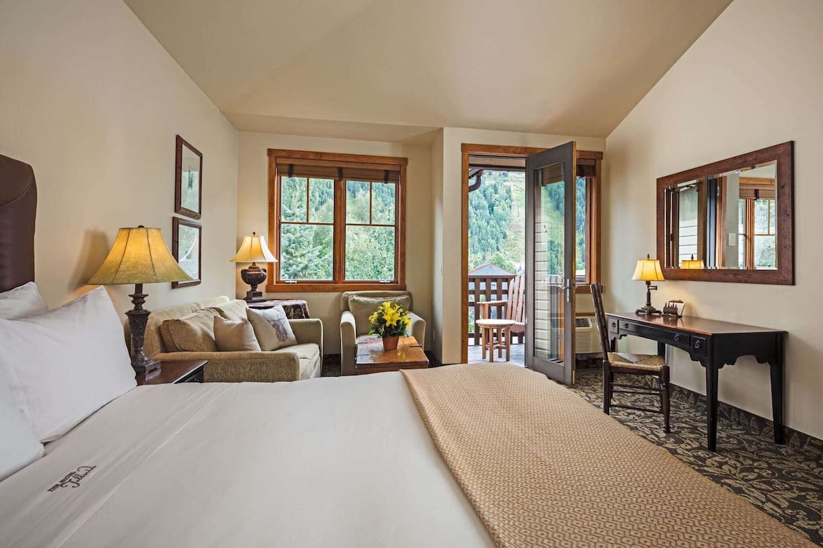 A hotel room with a bed, a desk, a chair, a sofa, and a balcony with a view of trees.