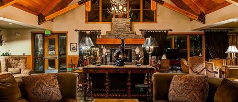 A spacious lobby with high ceilings, wooden beams, and a chandelier. There is a fireplace with antlers and a stone fireplace surround. The lobby has leather sofas and a coffee table with magazines.