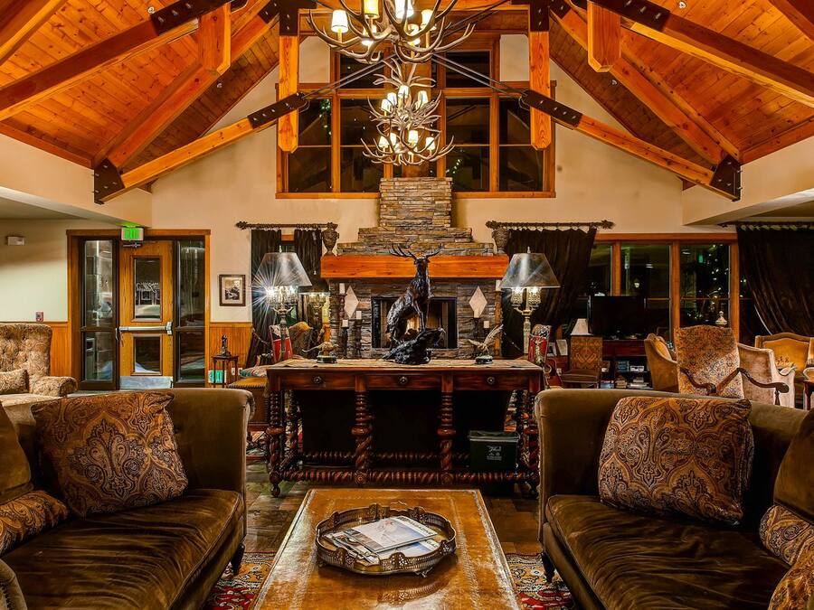 A spacious lobby with high ceilings, wooden beams, and a chandelier. There is a fireplace with antlers and a stone fireplace surround. The lobby has leather sofas and a coffee table with magazines.