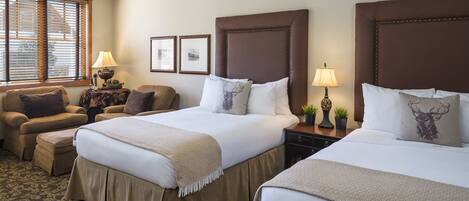 A hotel room with two beds, a sofa, a small table with lamps, framed pictures on the wall, and a window with blinds.
