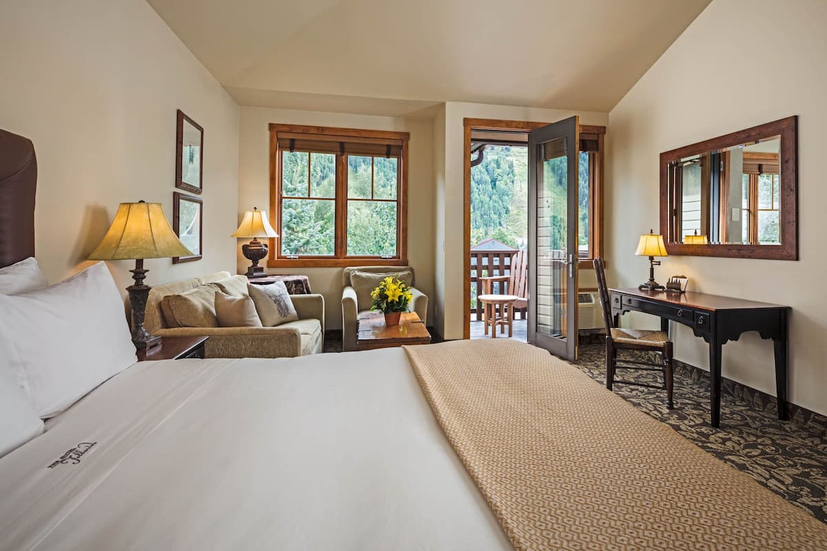 A hotel room with a bed, a desk, a chair, a sofa, and a balcony with a view of trees.