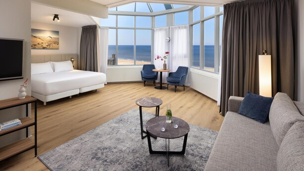 A modern hotel room with a large bed, a sofa, a small table, and a view of the sea through large windows. - NH Zandvoort (Zandvoort)