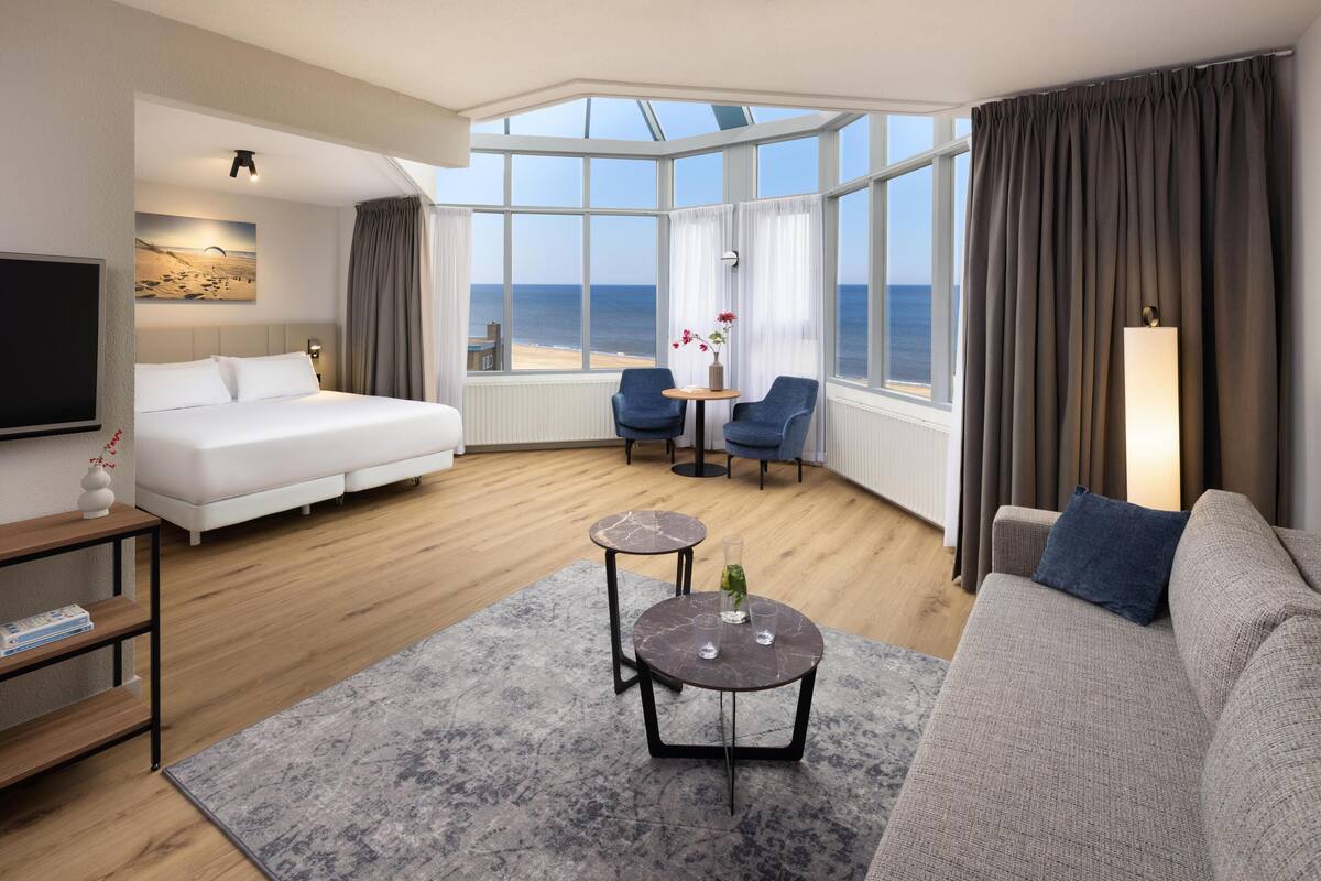 A modern hotel room with a large bed, a sofa, a small table, and a view of the sea through large windows.