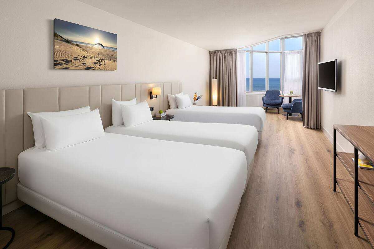 A hotel room with two beds, a desk, a chair, a TV, and a large window with a view of the sea.