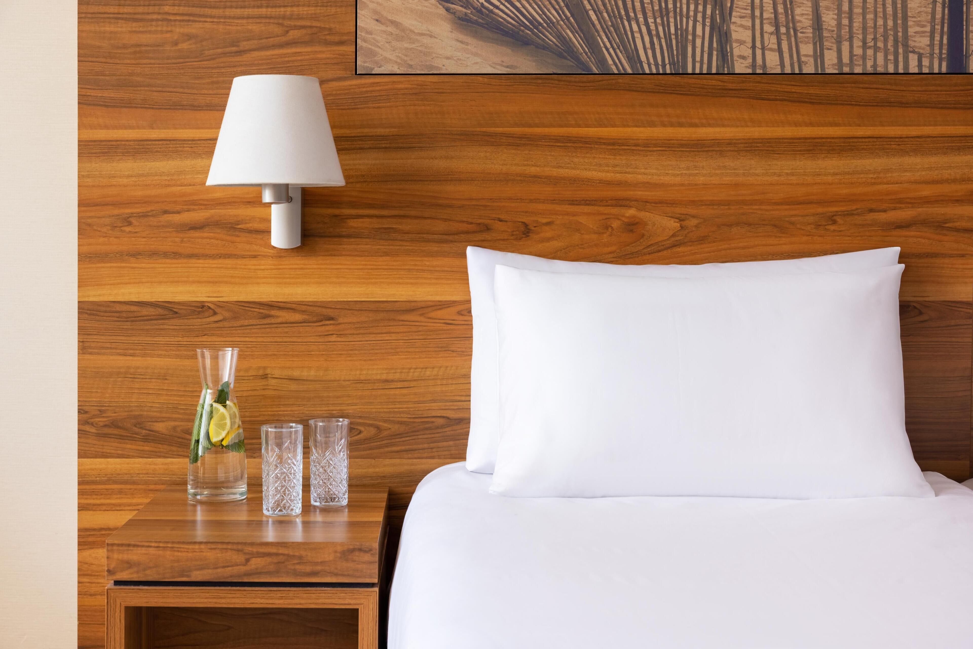 Una cama bien hecha con un almohadón blanco y un cabecero de madera. Una mesita de noche sostiene una lámpara blanca, un florero de vidrio con una rodaja de limón y dos vasos transparentes.