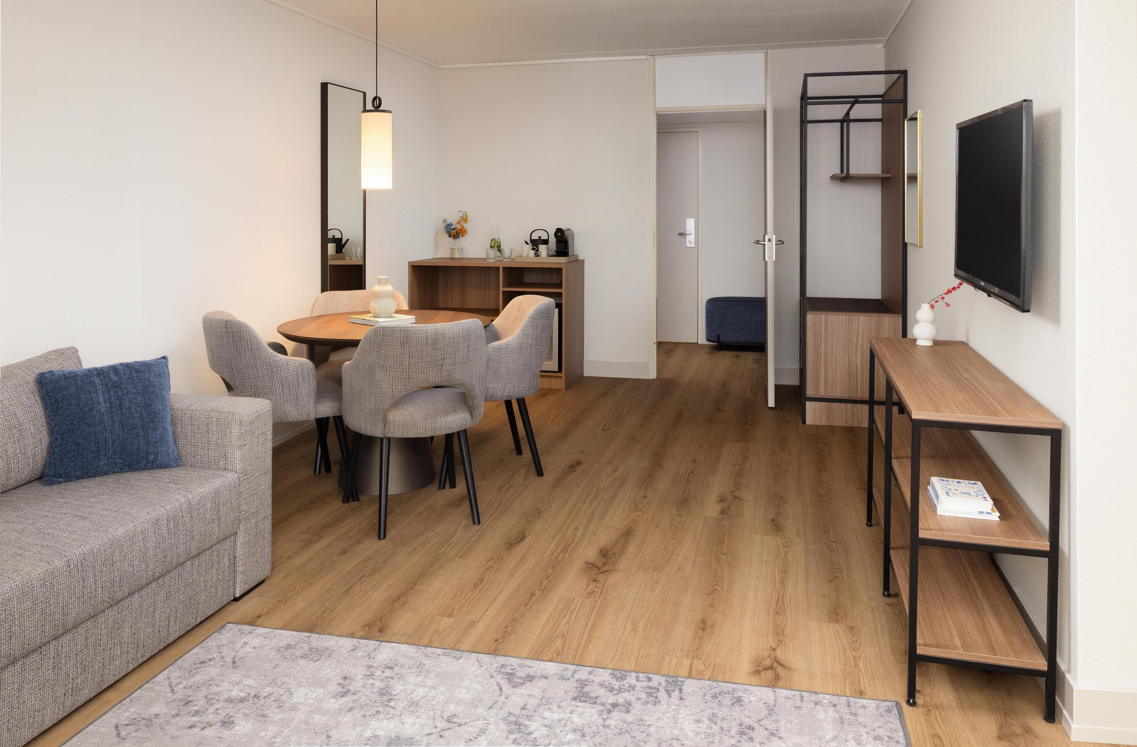 A modern living room with a sofa, dining table, and chairs, a TV mounted on the wall, and a bookshelf.