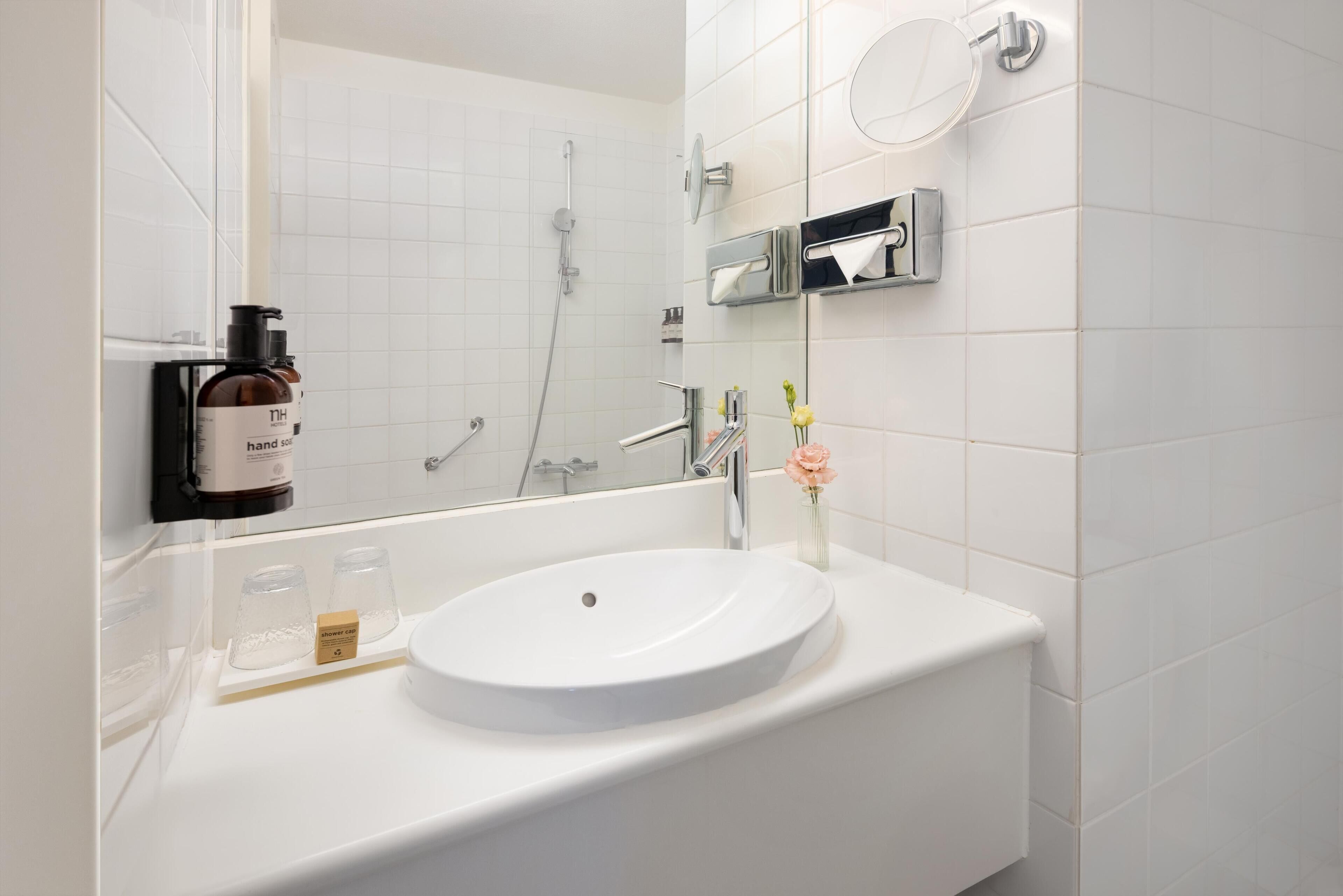 A modern bathroom with a white sink, a handheld showerhead, a towel dispenser, and a mirror.