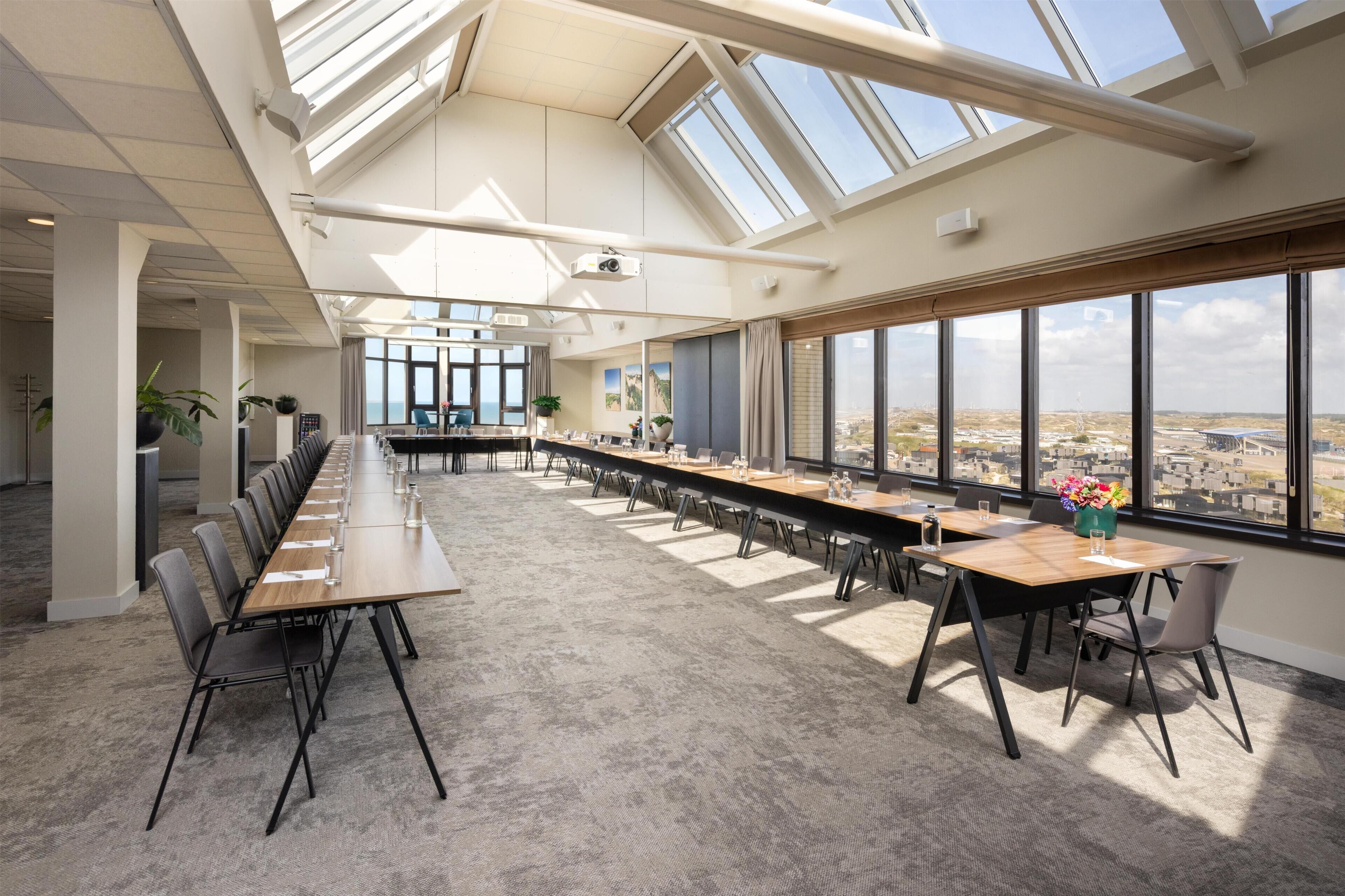 Un salón de conferencias con una larga mesa, sillas y grandes ventanales que ofrecen vistas de la ciudad.
