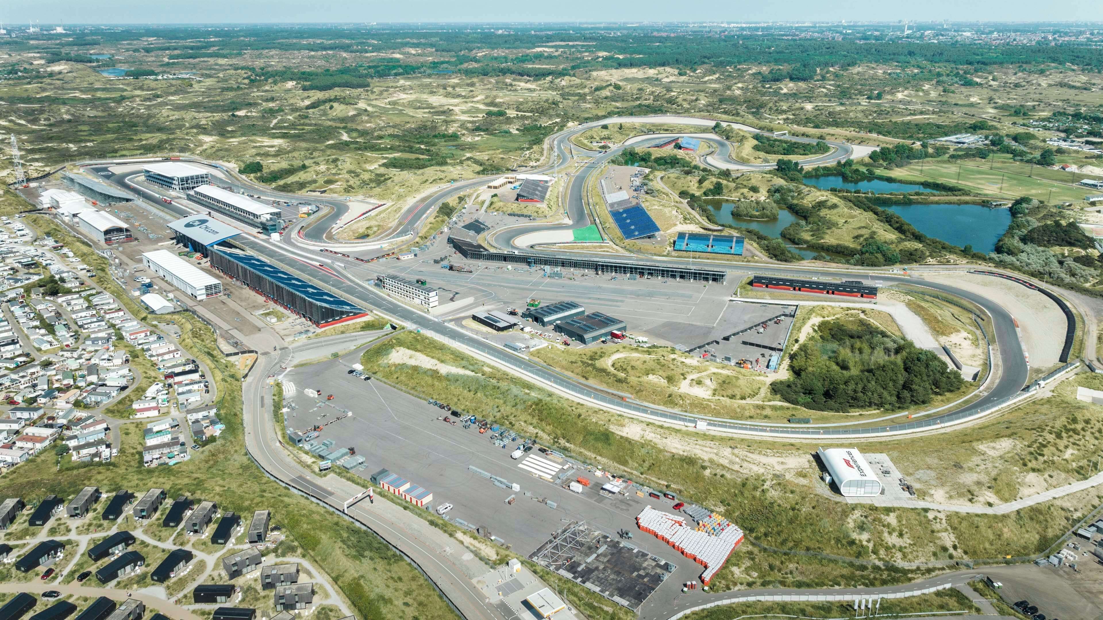 An aerial view of a racetrack with multiple turns, a large parking area, and a small lake.