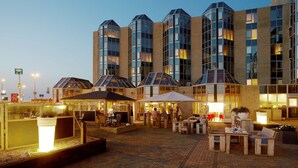 An outdoor patio area with tables and chairs, a bar area, and a building with multiple floors and large windows. - NH Zandvoort (Zandvoort)