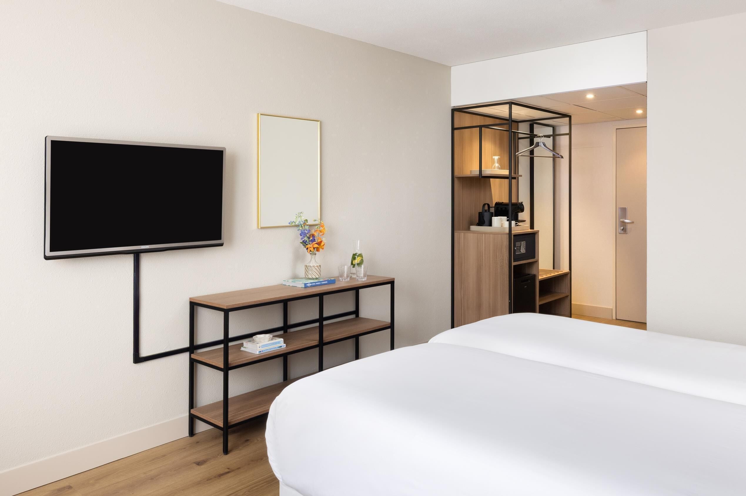 Ein modernes Hotelzimmer mit einem großen Bett, einem an der Wand montierten Fernseher, einem kleinen Konsolentisch mit Spiegel und einem begehbaren Kleiderschrank mit Kleiderhängeorganizer.