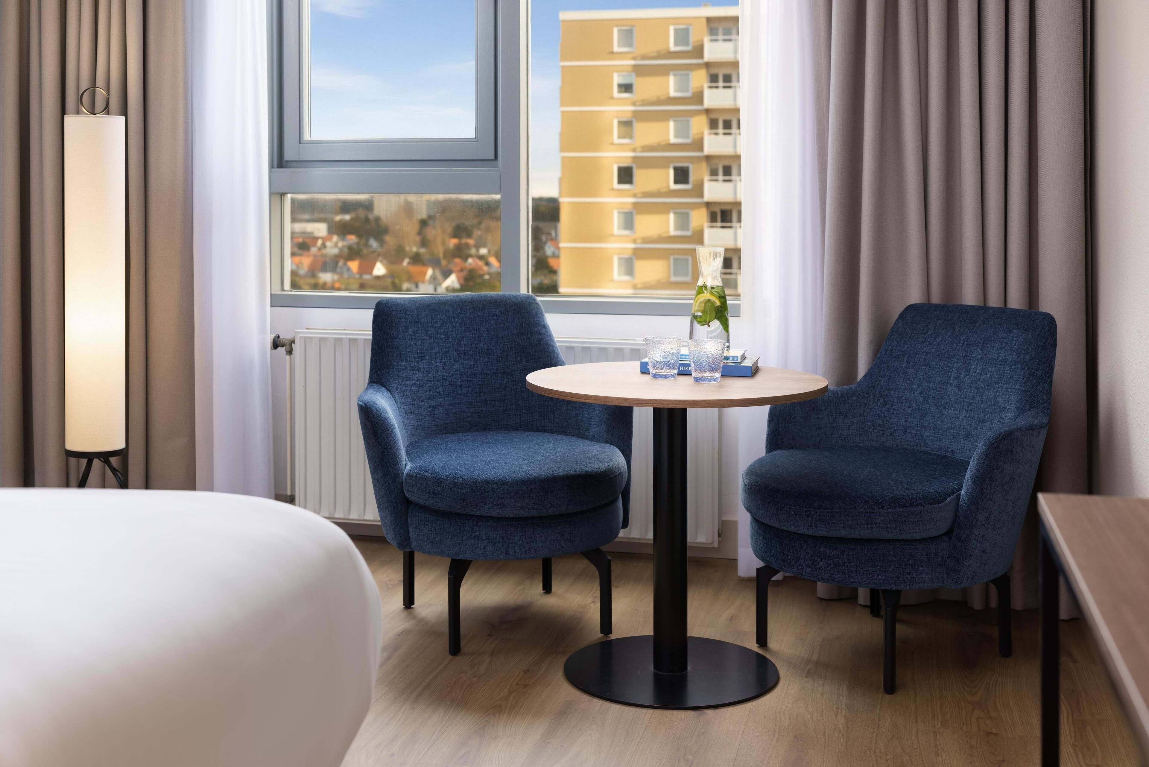 Ein Hotelzimmer mit zwei blauen Sesseln um einen kleinen runden Tisch, einer Lampe und Blick auf ein Gebäude durch ein Fenster.