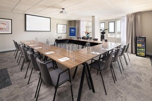 A conference room with a long table, chairs, a projector, a screen, a beverage cooler, and framed artwork on the walls. - NH Zandvoort (Zandvoort)