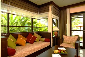 A room with a sofa and a chair, a table with fruit, and a view of greenery outside.