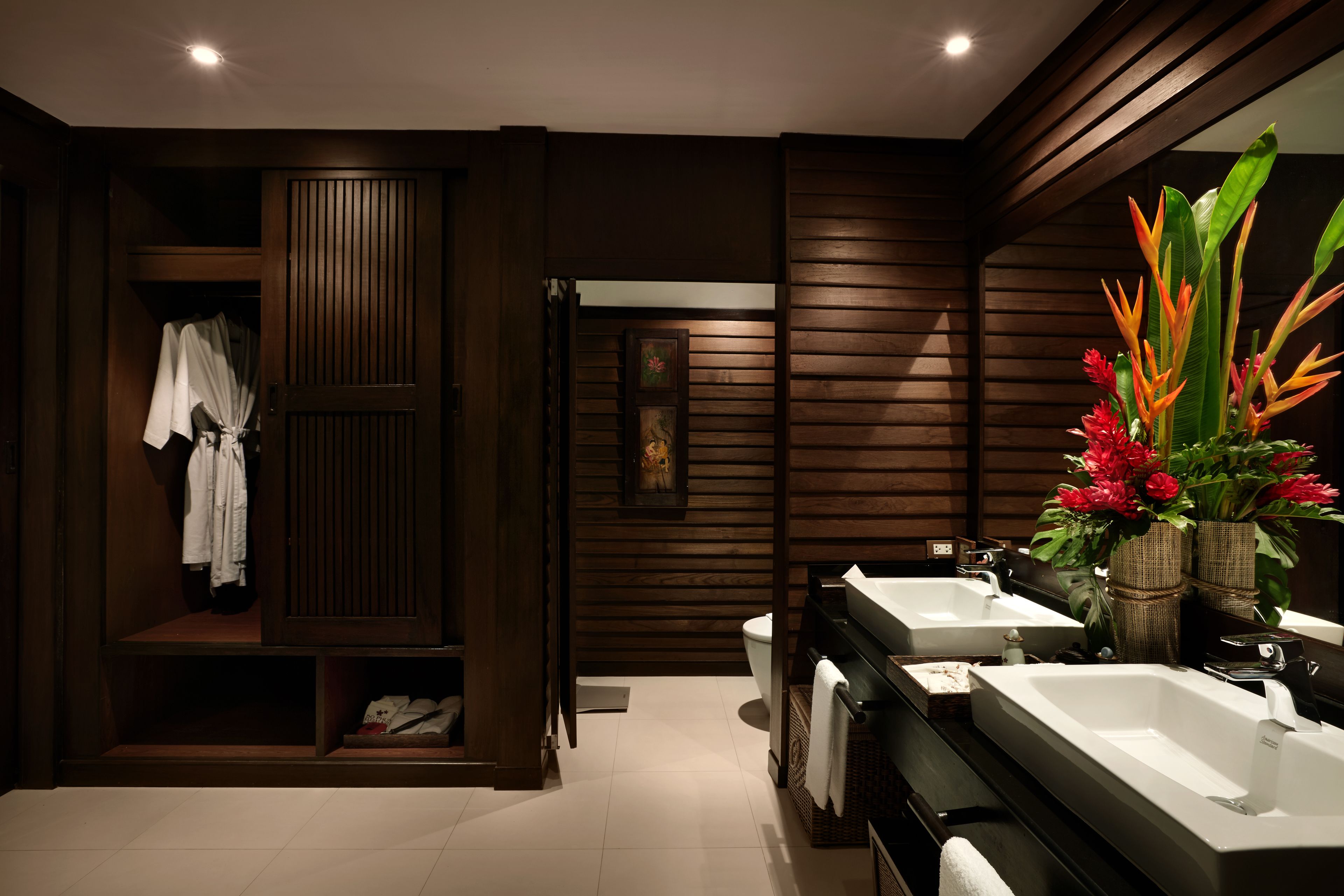 A modern bathroom with two sinks, a robe closet, and a mirror reflecting a vase of flowers.