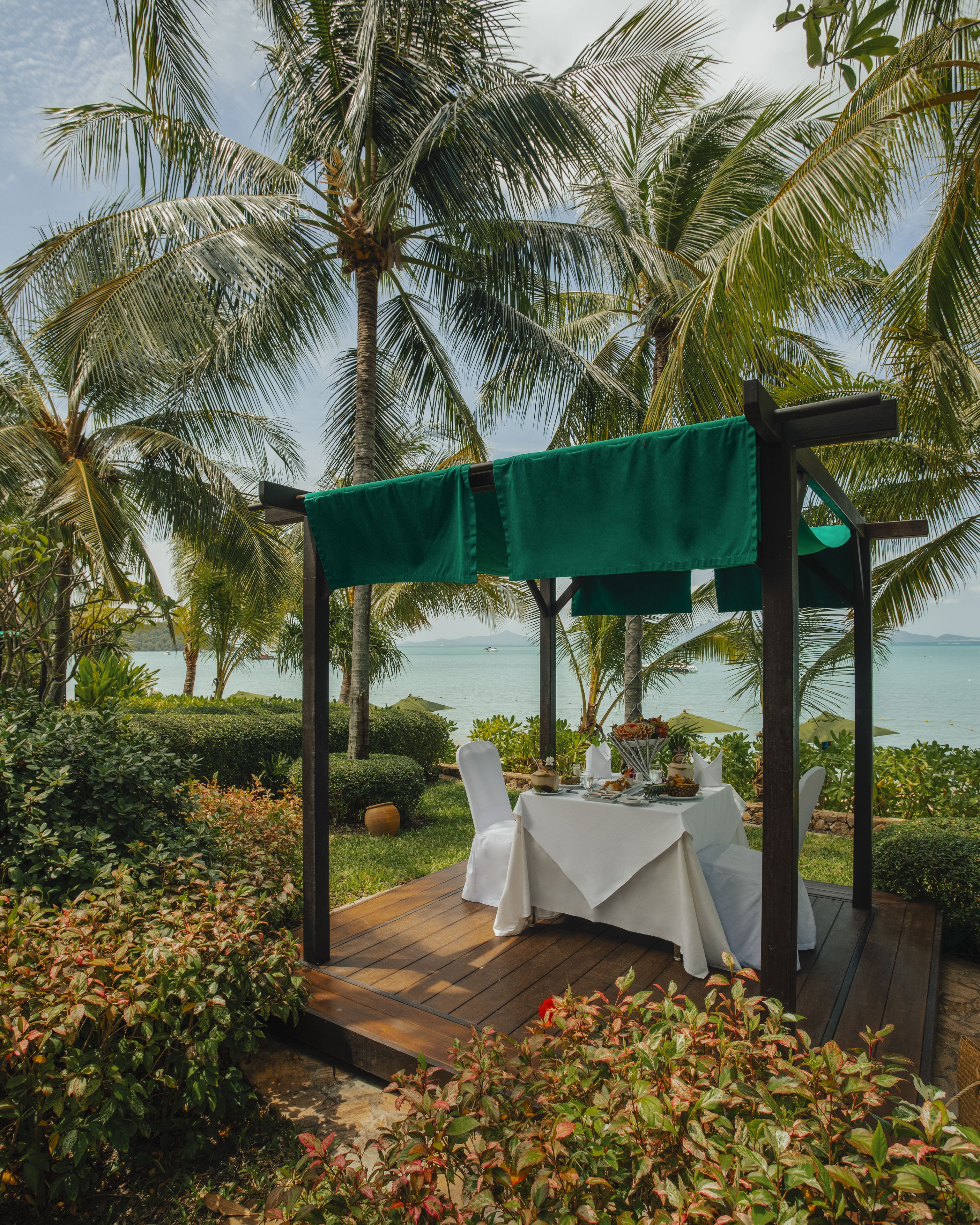 An outdoor dining area with a wooden deck, white tablecloths, and chairs, surrounded by lush greenery and palm trees, overlooking a body of water.