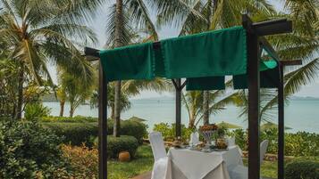 An outdoor dining area with a wooden deck, white tablecloths, and chairs, surrounded by lush greenery and palm trees, overlooking a body of water.