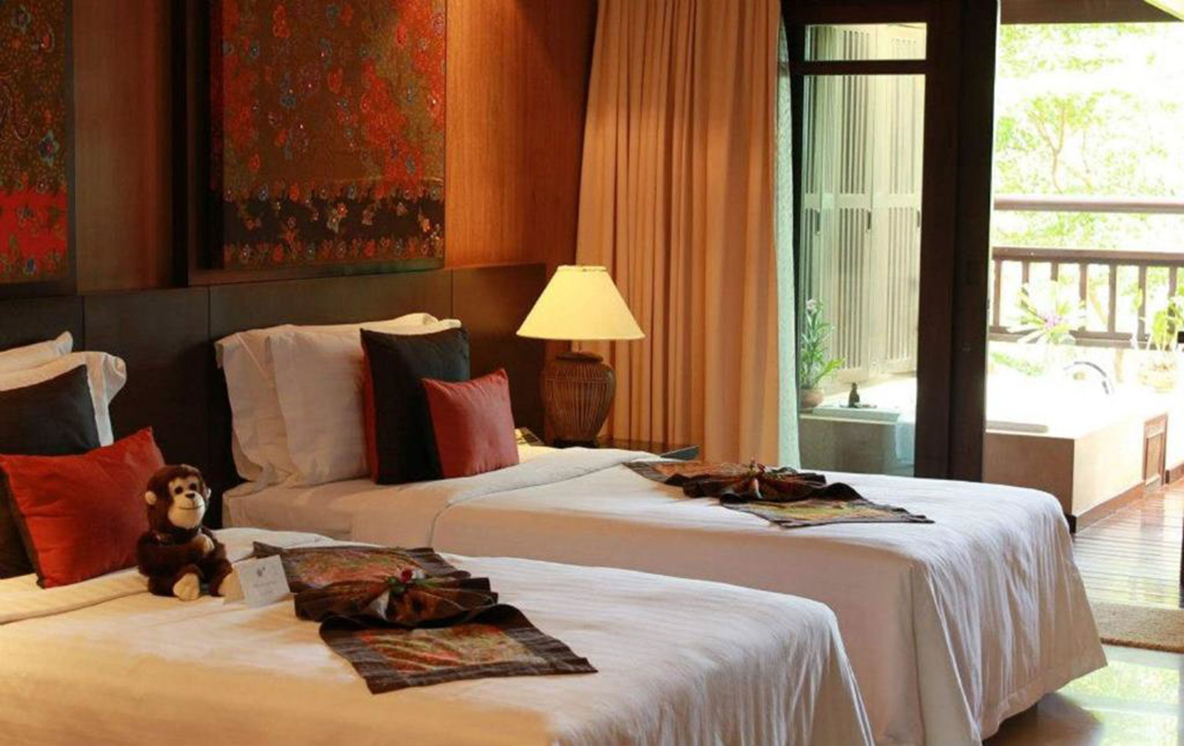 Een hotelkamer met twee bedden, een nachtkastje met een lamp, een pluche speeltje en een raam met uitzicht op groen.