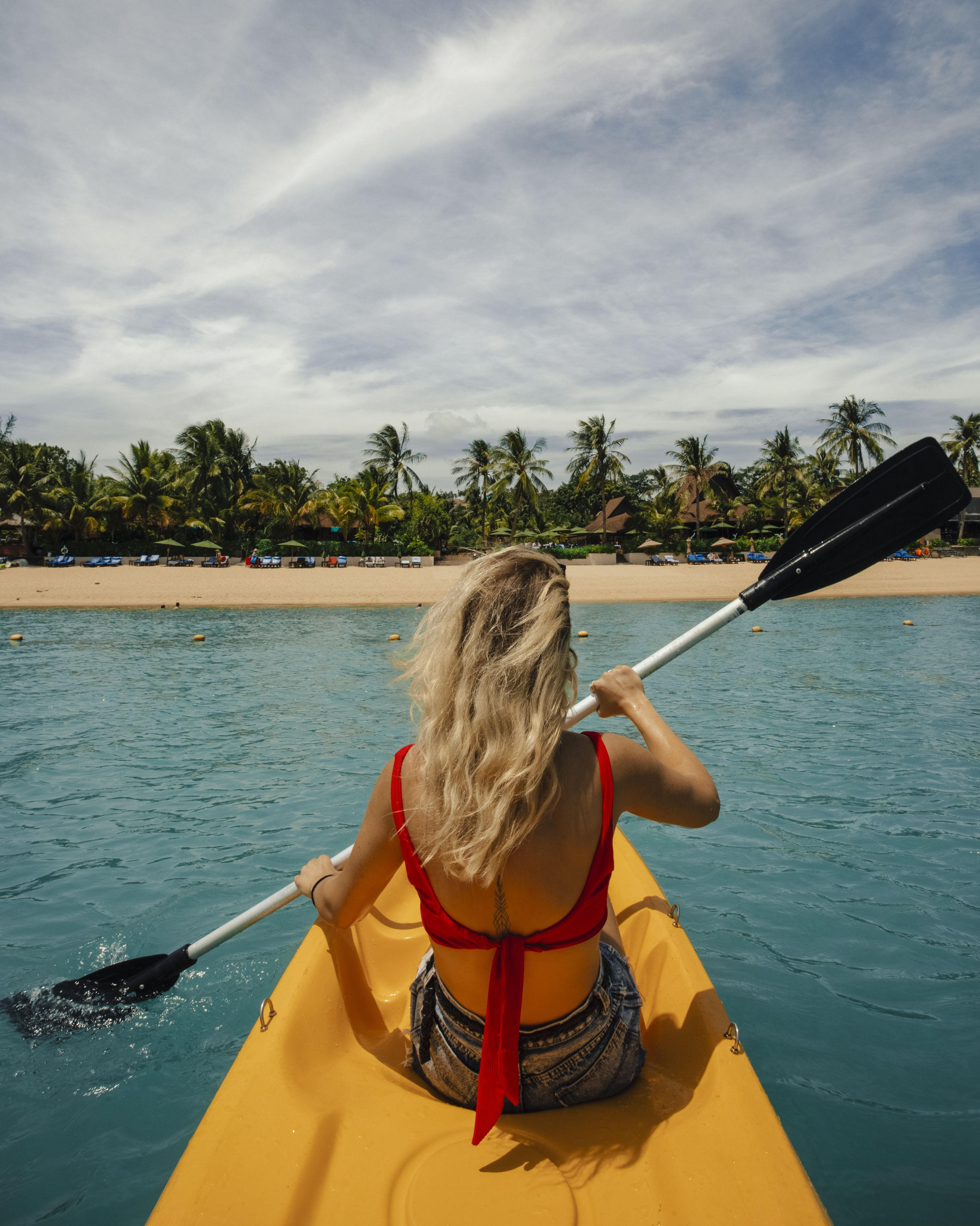 A person in a yellow kayak on a calm sea with a beach and palm trees in the background.