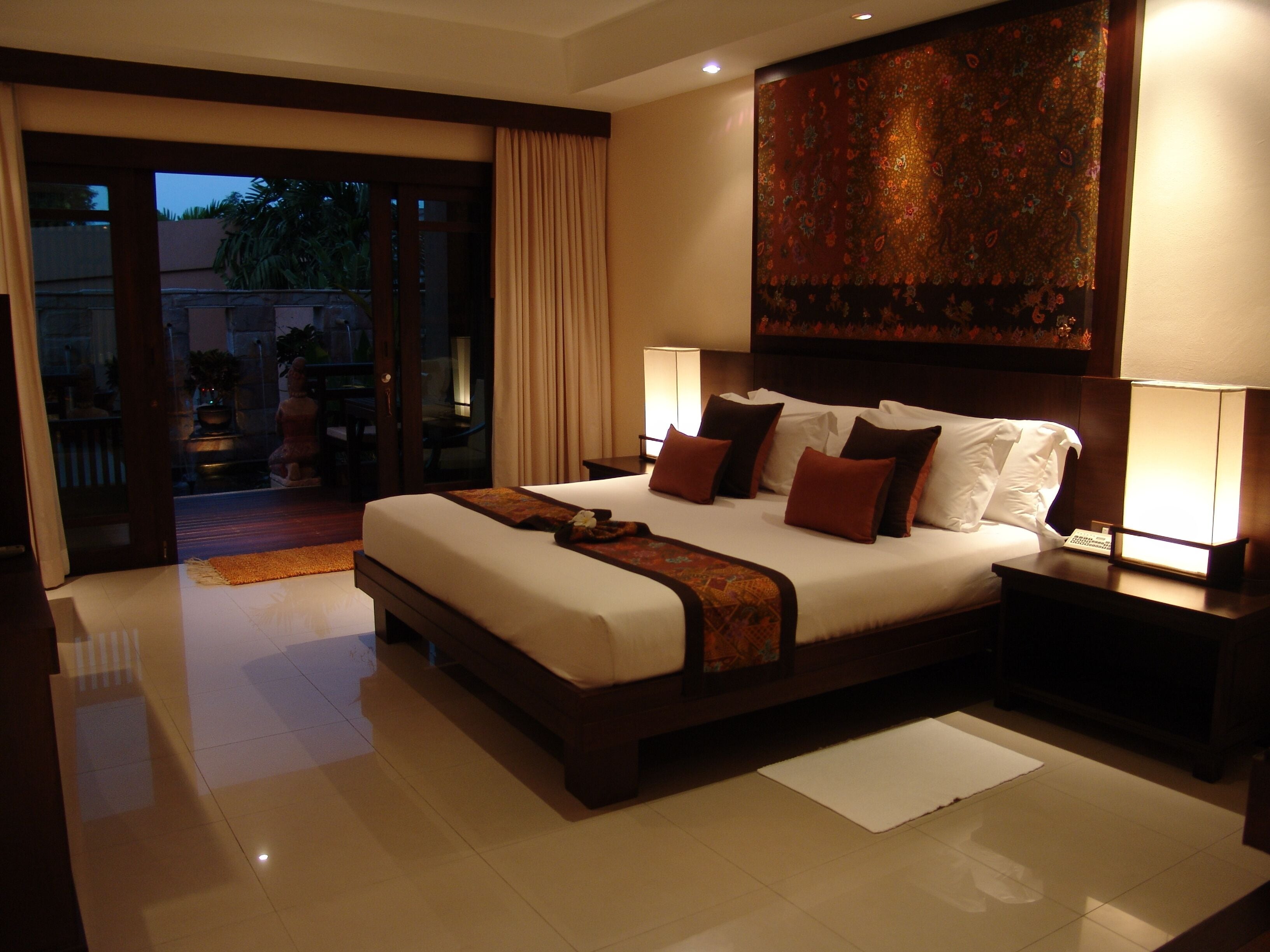 A spacious hotel bedroom with a large bed, two bedside lamps, and a view through the glass door.