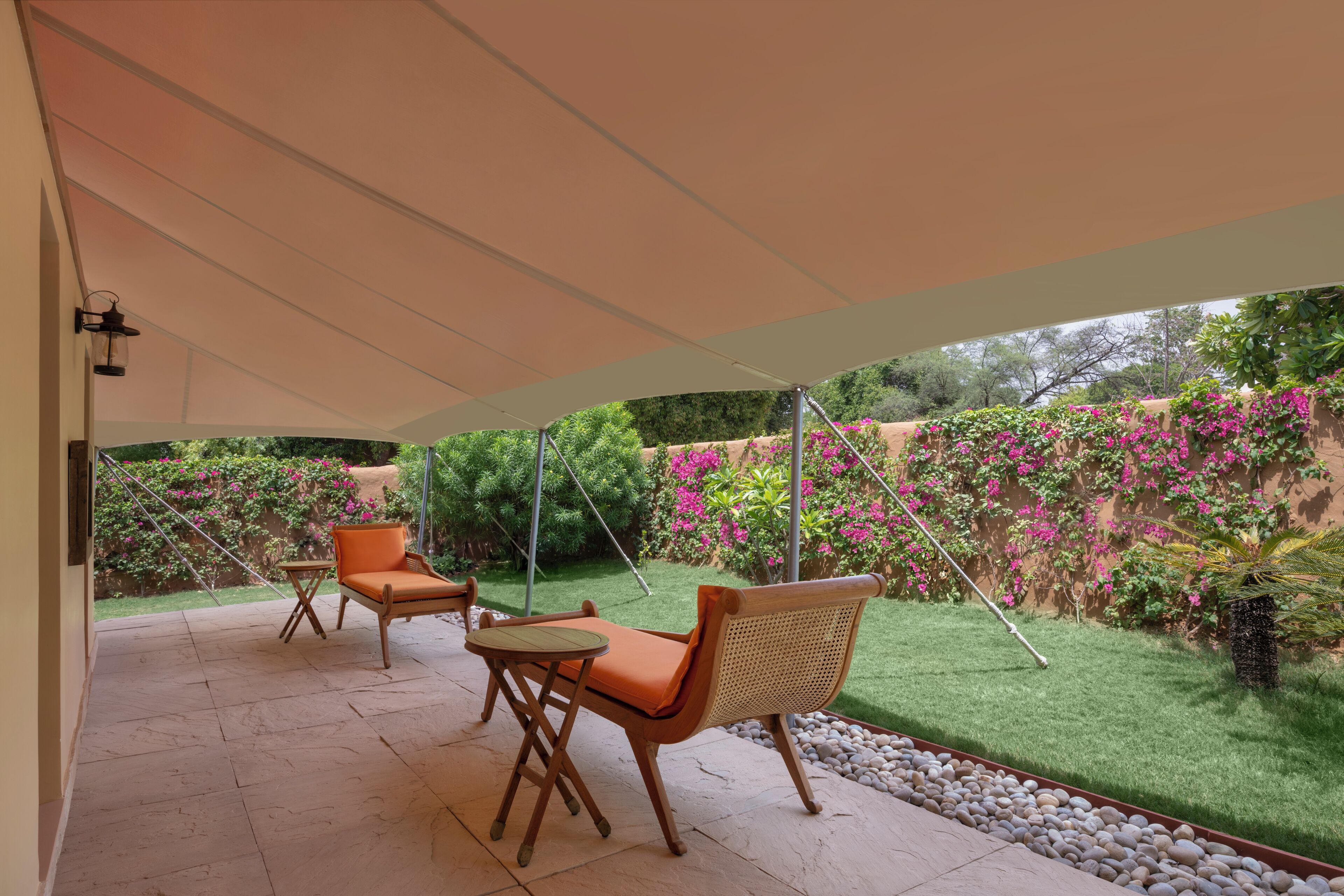 Un patio al aire libre con dos sillas de madera y una mesa pequeña, un dosel blanco sobre la cabeza, y vistas de un jardín con flores rosas y follaje.