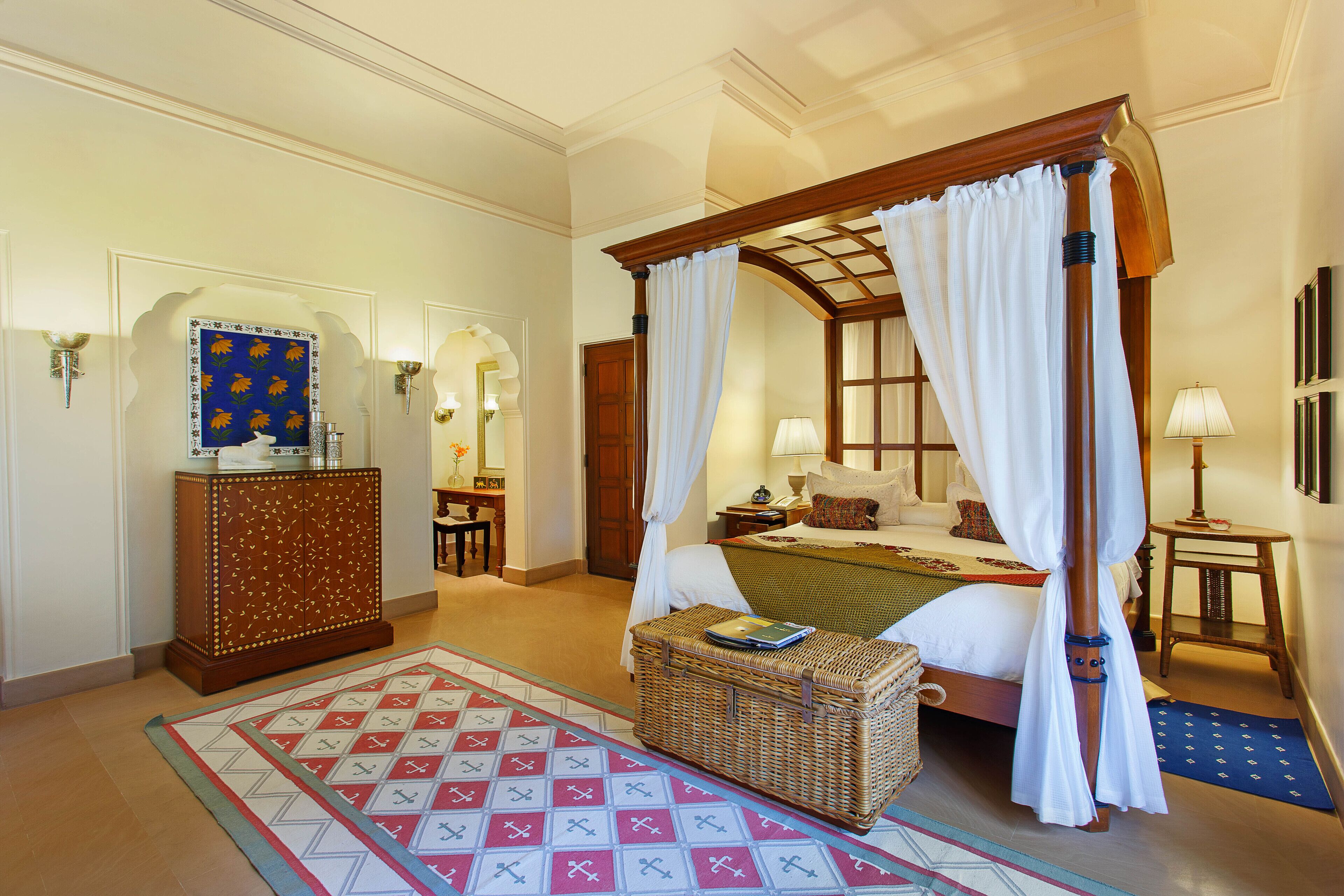 Uma cama com dossel e colunas, cabeceira de madeira, mesa de cabeceira de vime, tapete estampado e um espelho decorativo na parede.