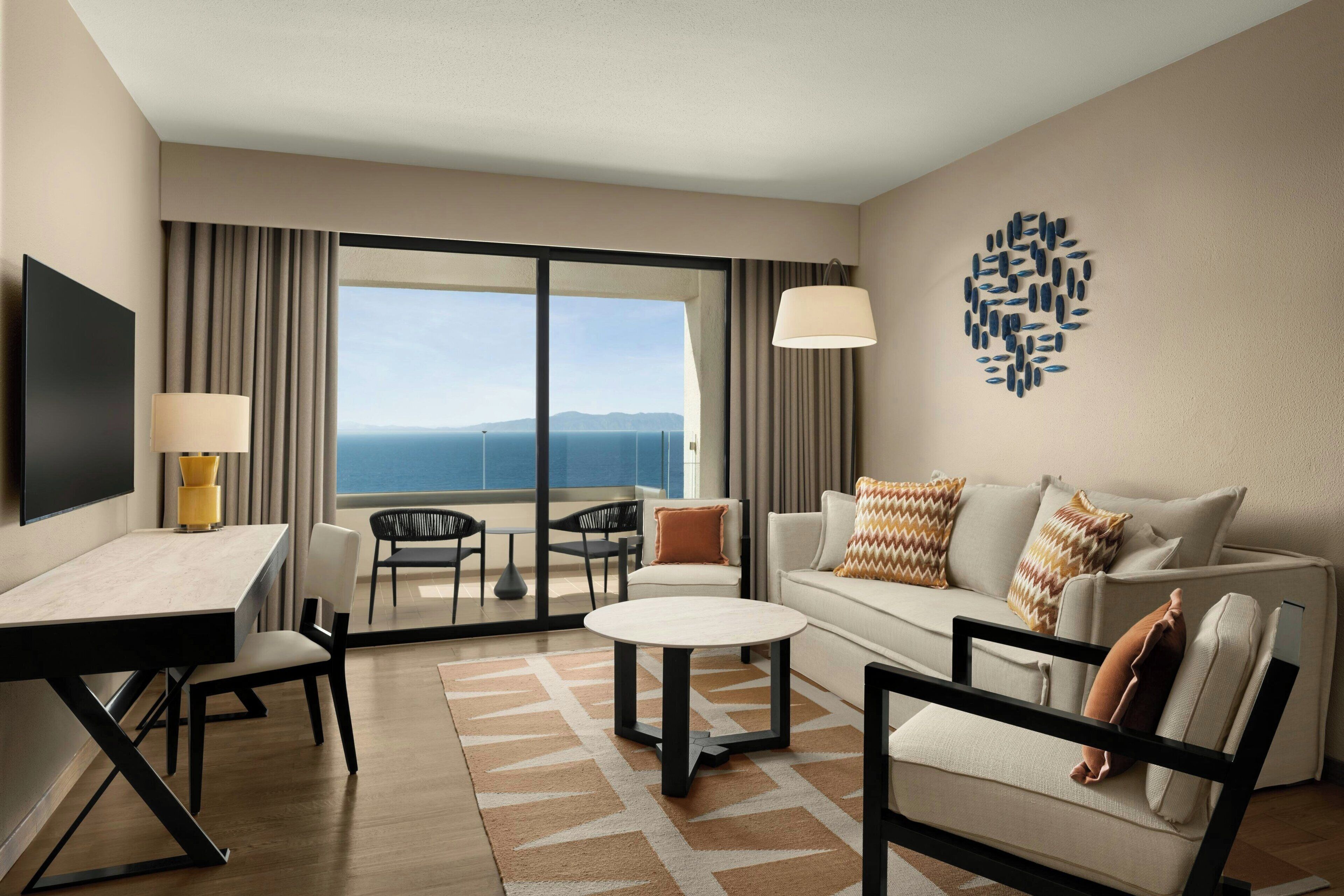 A hotel room with a flat-screen TV, a dining table, a sofa, a coffee table, and a view of the ocean.
